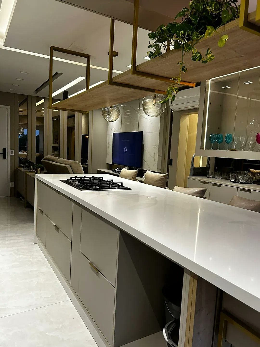 Cozinha planejada com ilha, bancada branca e prateleira suspensa dourada com plantas – projeto real Estillo Design Zona Leste SP